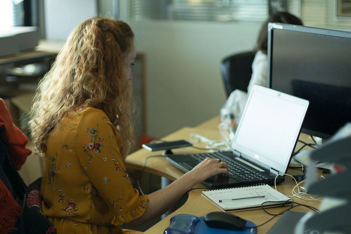 Une femme travaille sur un ordinateur portable à un bureau, avec un carnet de notes à ses côtés.